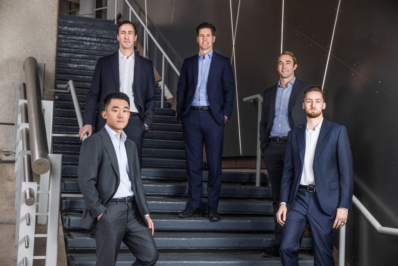 Five businessmen in suits posing on stairs in a modern office setting.